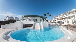 Un área de piscina moderna con diseño circular, un puente de cristal y un edificio blanco con balcones.