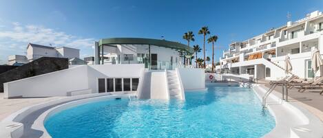 Un área de piscina moderna con diseño circular, un puente de cristal y un edificio blanco con balcones.