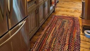 A modern kitchen with wooden cabinets, a stainless steel refrigerator, hanging light bulbs, and a colorful woven rug.