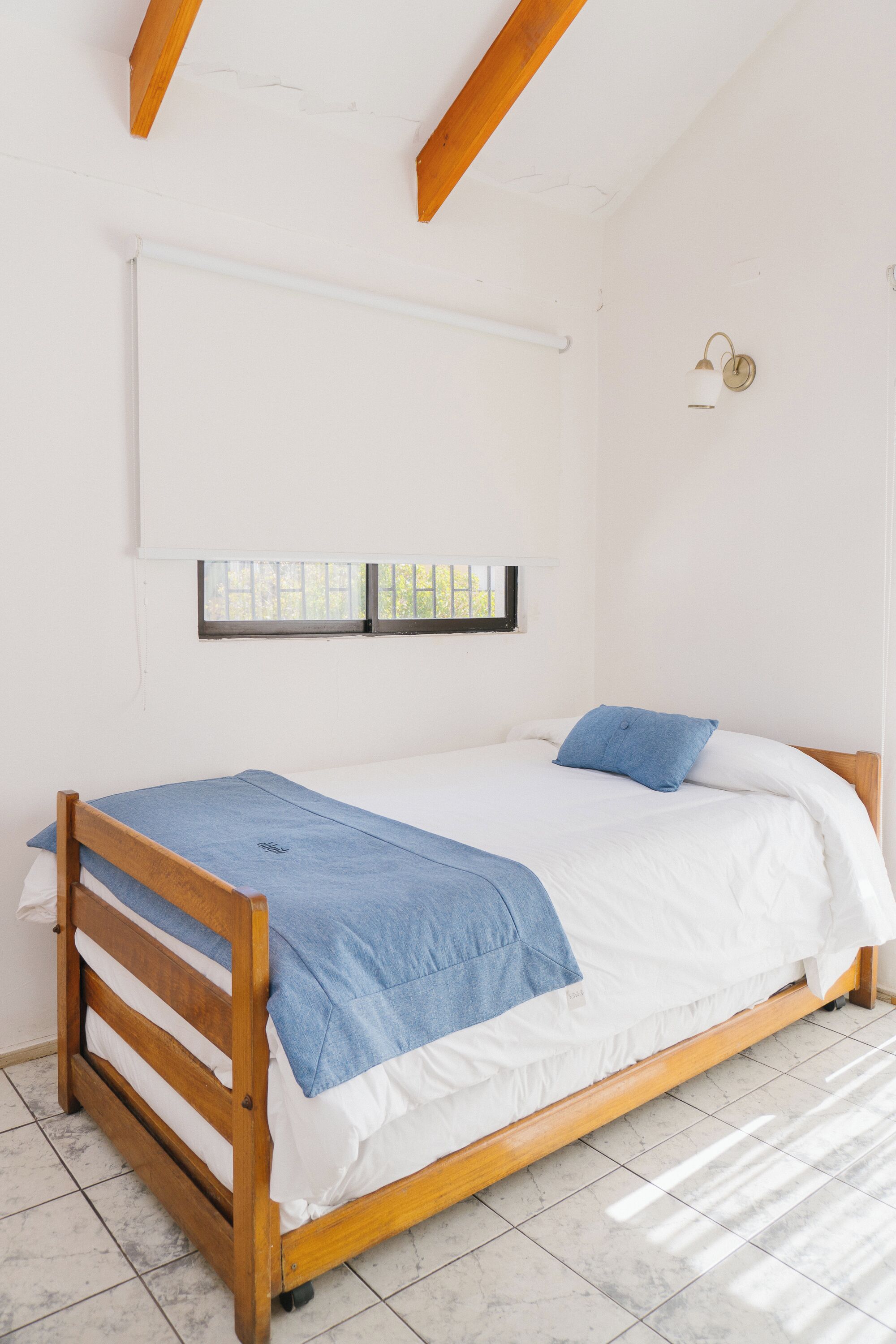 A single bed with a wooden frame and a blue blanket, white bedding, a window with bars, and a white wall with a light fixture.