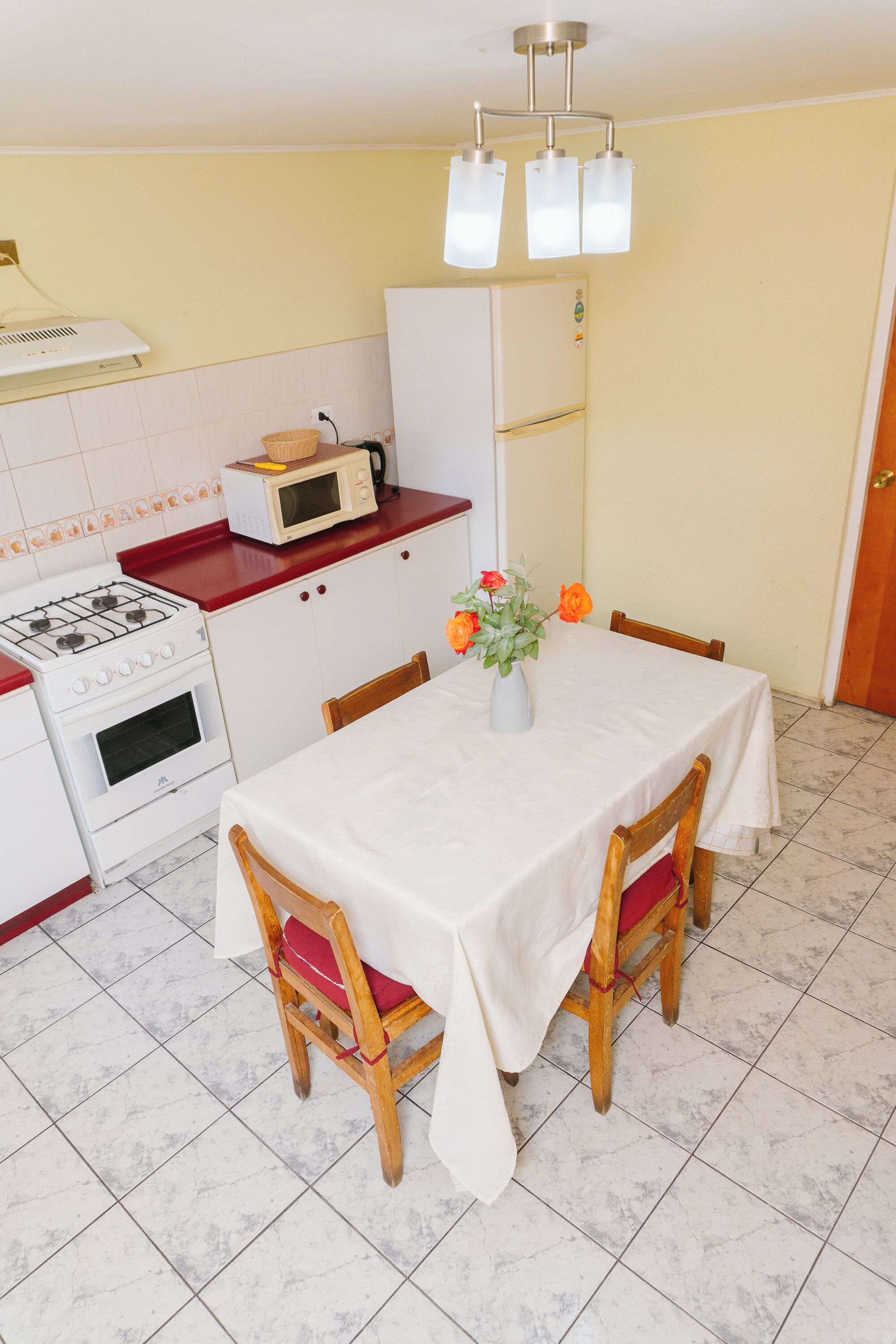 A compact kitchen with a stove, microwave, and refrigerator. There is a dining table with chairs and a vase with flowers.