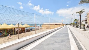 Un paseo marítimo con una zona de playa arenosa, un cerramiento de malla y una hilera de palmeras.