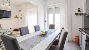 Un comedor moderno con una mesa larga, sillas grises y un cuenco de fruta. Se observa también una cocina con estufa y fregadero.