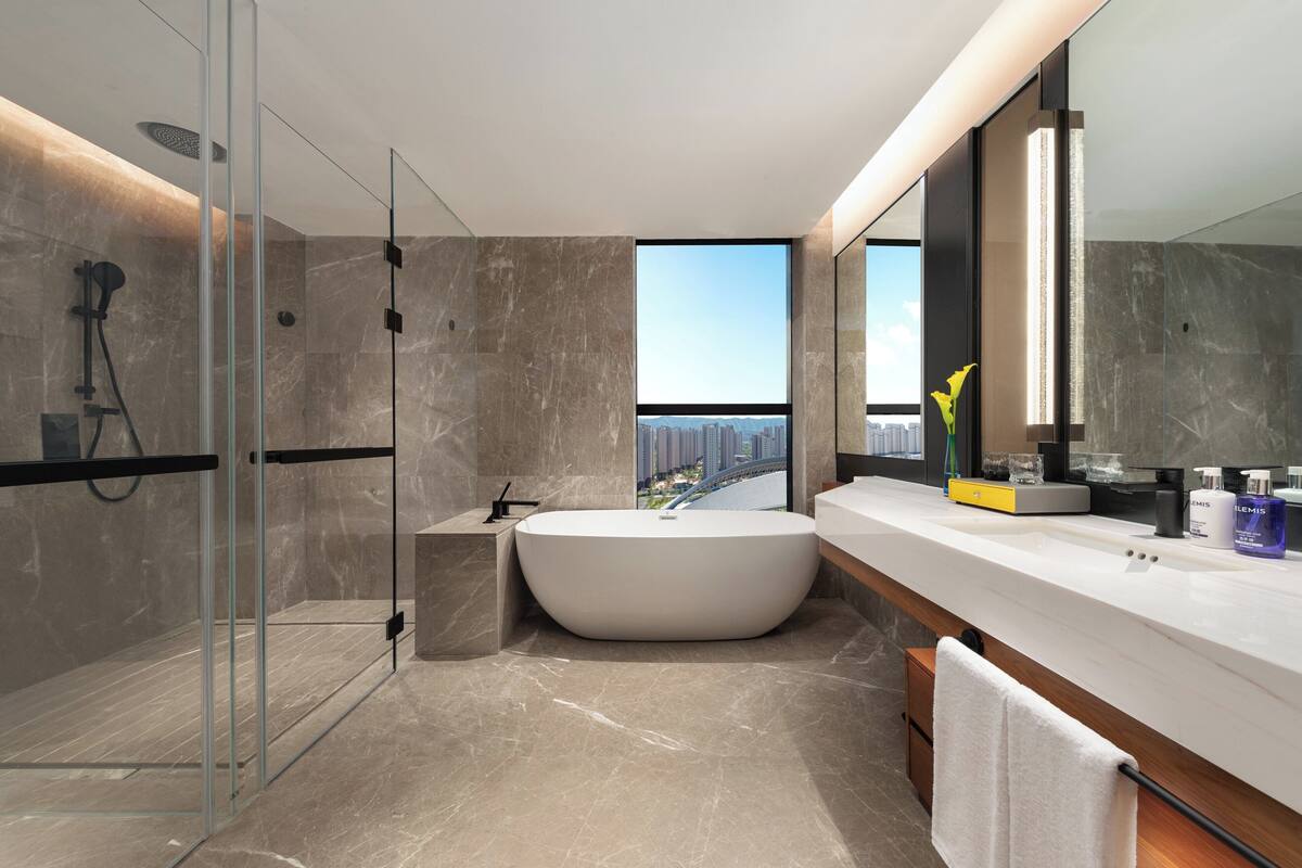 Un baño moderno con una bañera grande, una ducha con cerramiento de vidrio y vistas a un paisaje urbano a través de un ventanal.