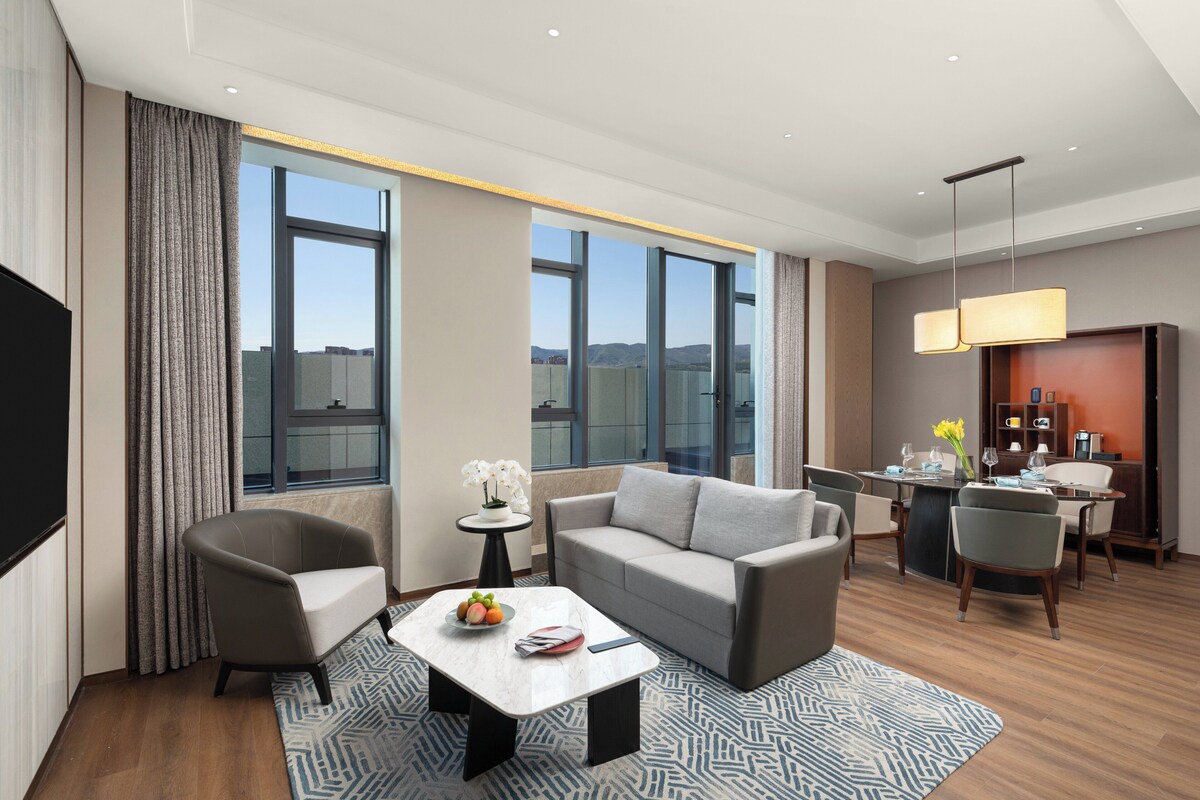 A modern hotel room with a sofa, armchairs, a dining table, and a TV.