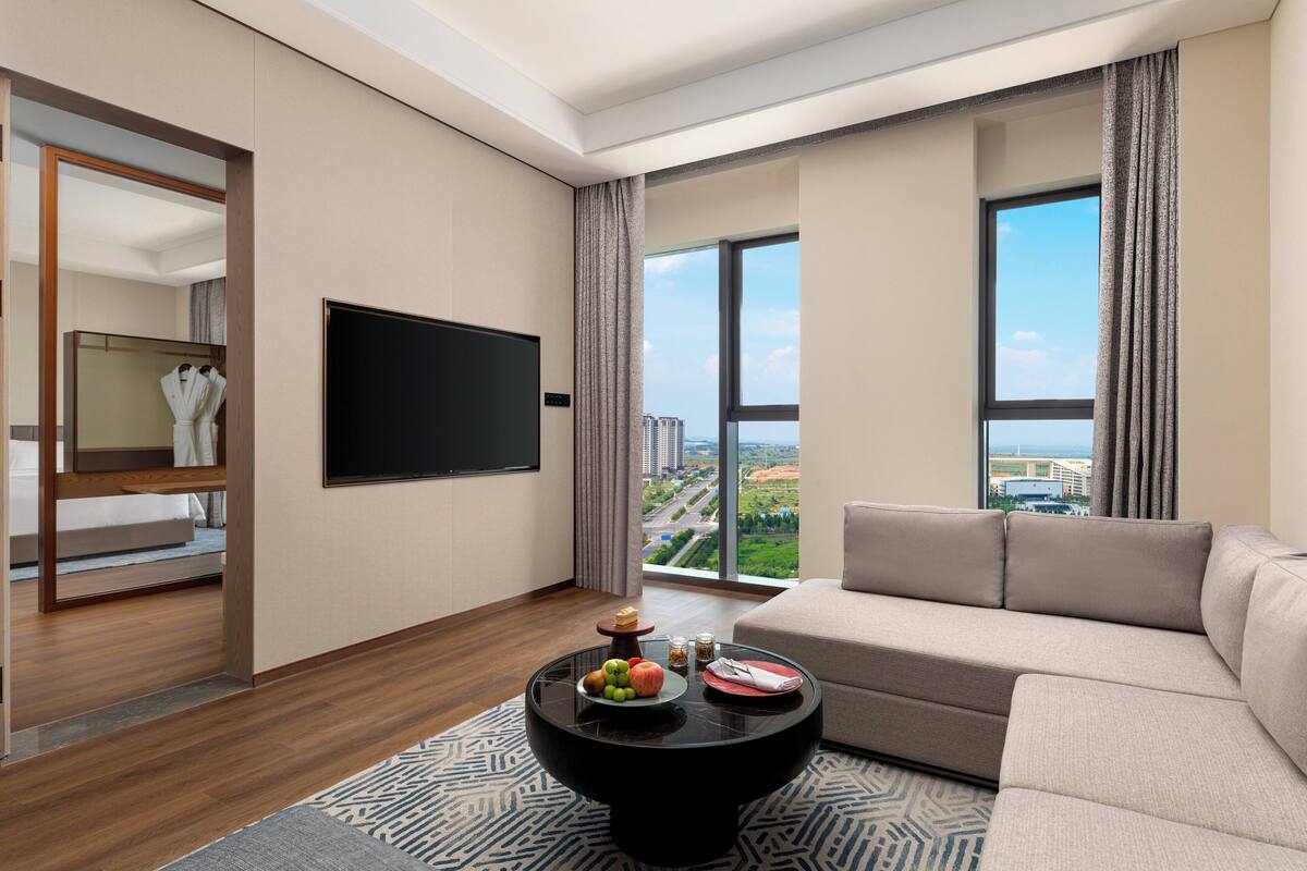 Un salón moderno con un ventanal grande, un televisor de pantalla plana, un sofá y una mesa de centro con frutas y aperitivos.