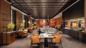 Un restaurante moderno con una zona de comedor central que presenta una mesa redonda, rodeada de sillas color naranja. Hay varias mesas de comedor con sillas, y una zona de bar con una barra y taburetes. El techo tiene un diseño de cuadrícula y hay lámparas colgantes.