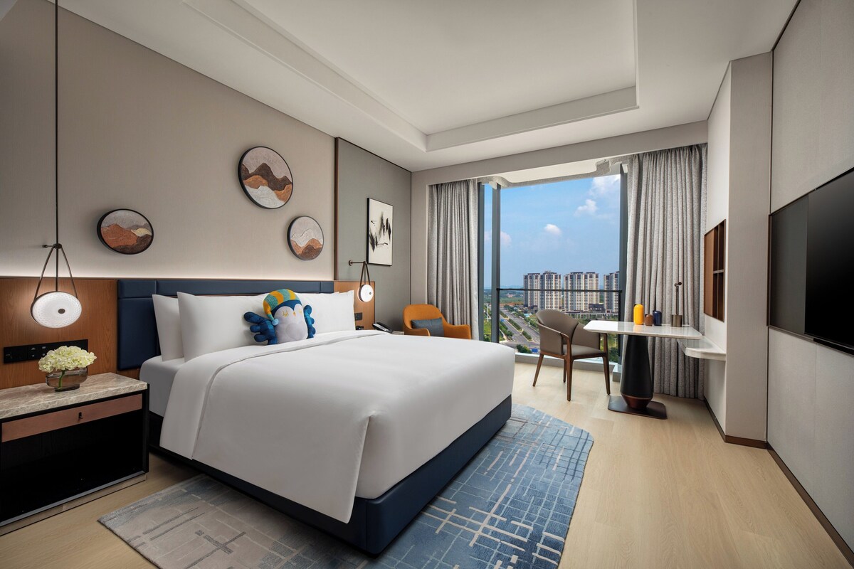 A modern hotel room with a large bed, a desk, a chair, a TV, and a view of the city.