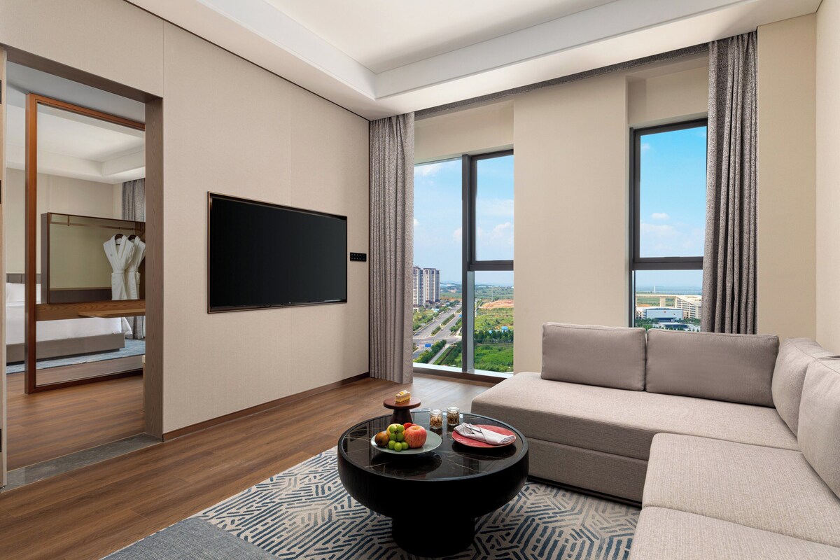 A modern living room with a large window, a flat-screen TV, a sofa, and a coffee table with fruits and snacks.