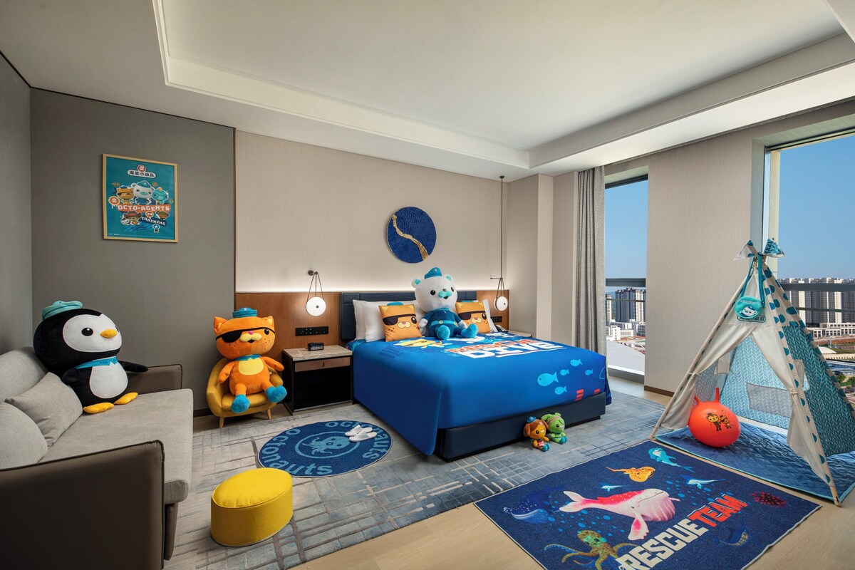 A hotel room with a bed, a sofa, a play tent, and a large window offering a city view.