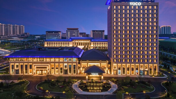Un hotel de gran altura con un llamativo letrero 'YOCO', que presenta un diseño arquitectónico moderno, una terraza en la azotea y un jardín paisajístico.
