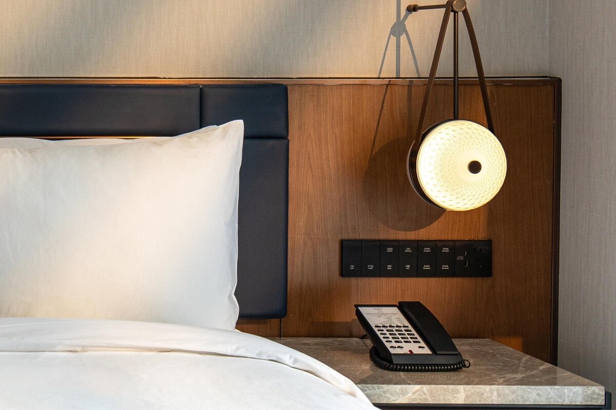 A hotel room with a bed, a bedside table with a telephone, and a wall-mounted light fixture.