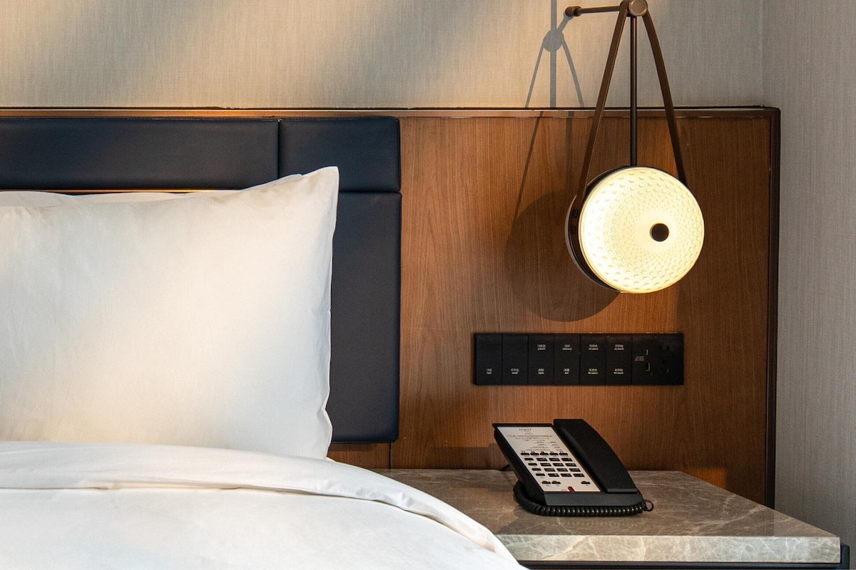 Una habitación de hotel con una cama, una mesita de noche con teléfono y una lámpara fijada a la pared.