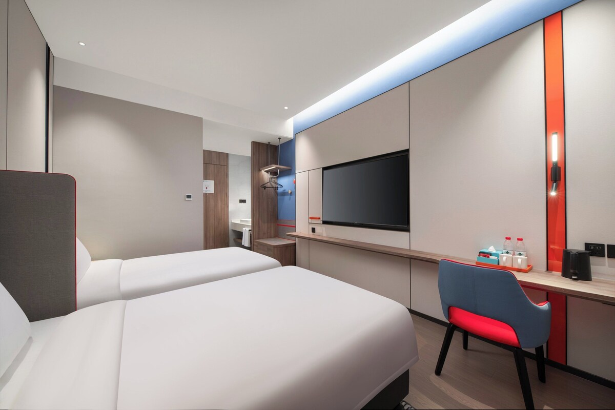 Habitació d'hotel moderna amb dos llits, una escriptori amb un televisor, una cadira i una taula petita amb ampolles.