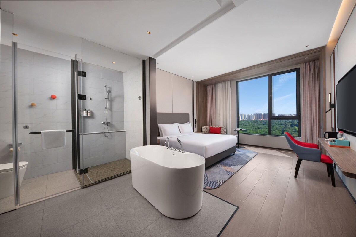 Habitació d'hotel moderna amb dutxa envidriada, banyera gran, llit amb vista a la ciutat i escriptori amb cadira.