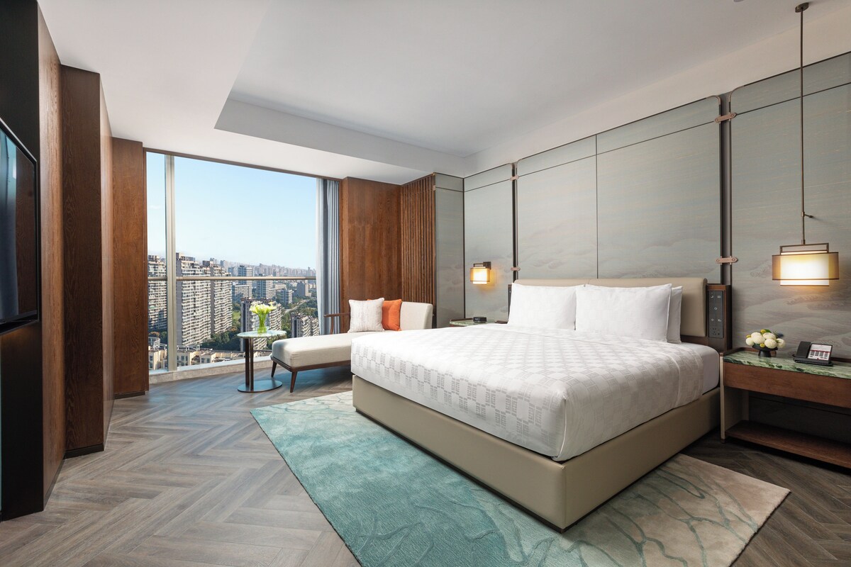 A modern hotel room with a large bed, a seating area, a TV, and a city view.