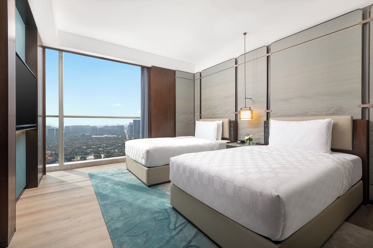 A modern hotel room with a large window, two beds, a flat-screen TV, and a city view.