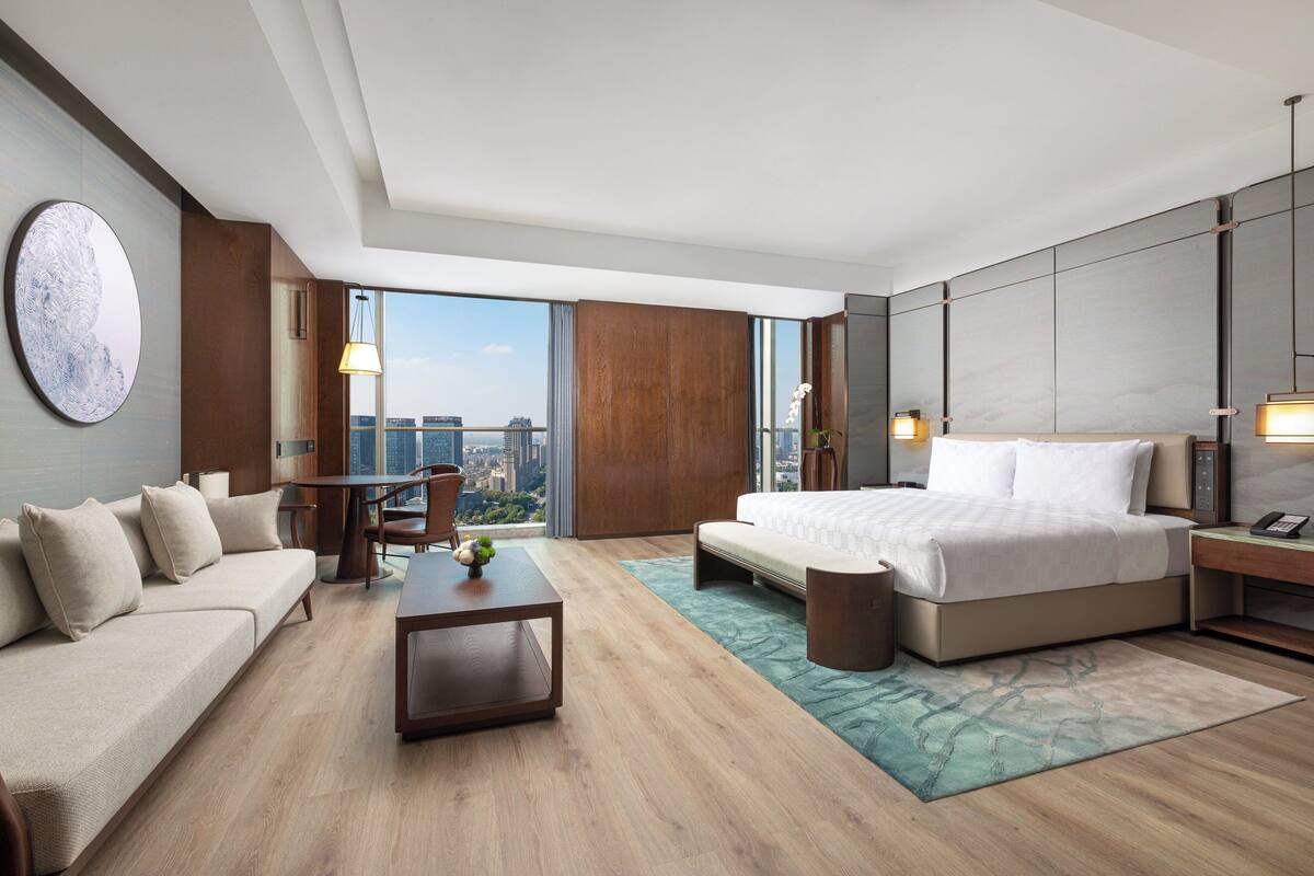 A modern hotel room with a large bed, a sofa, a dining table, and a view of the city.