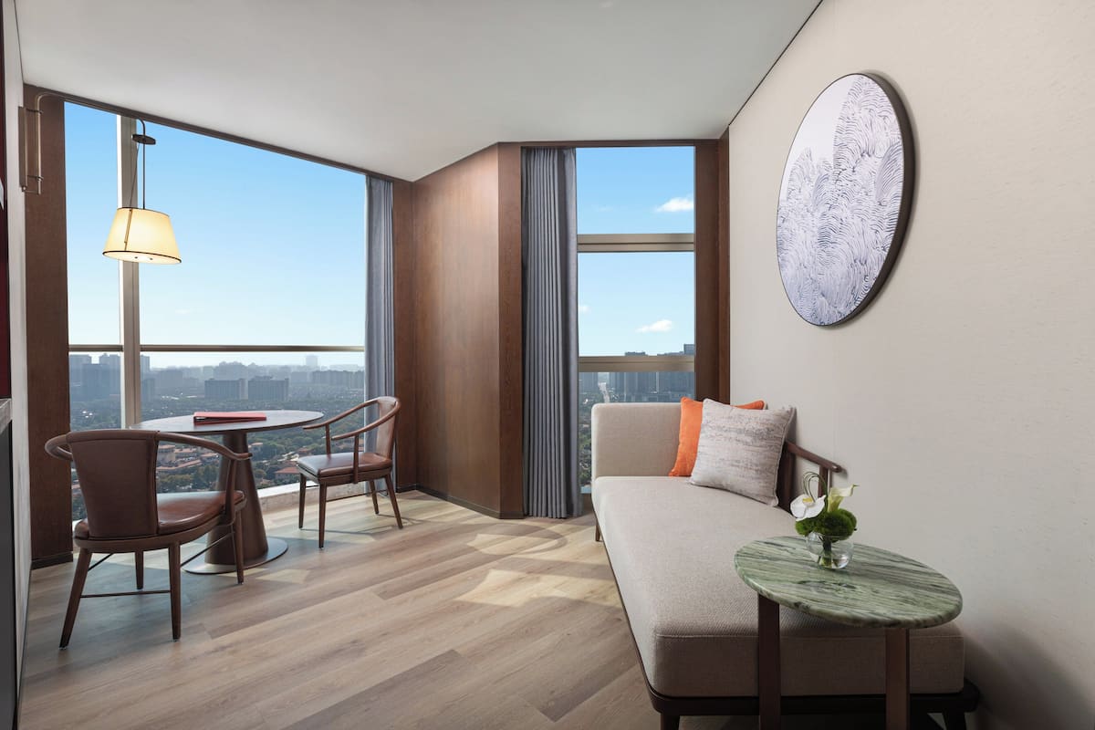 A modern hotel room with a sofa, a small table with a vase, a dining area with chairs, and large windows offering a city view.