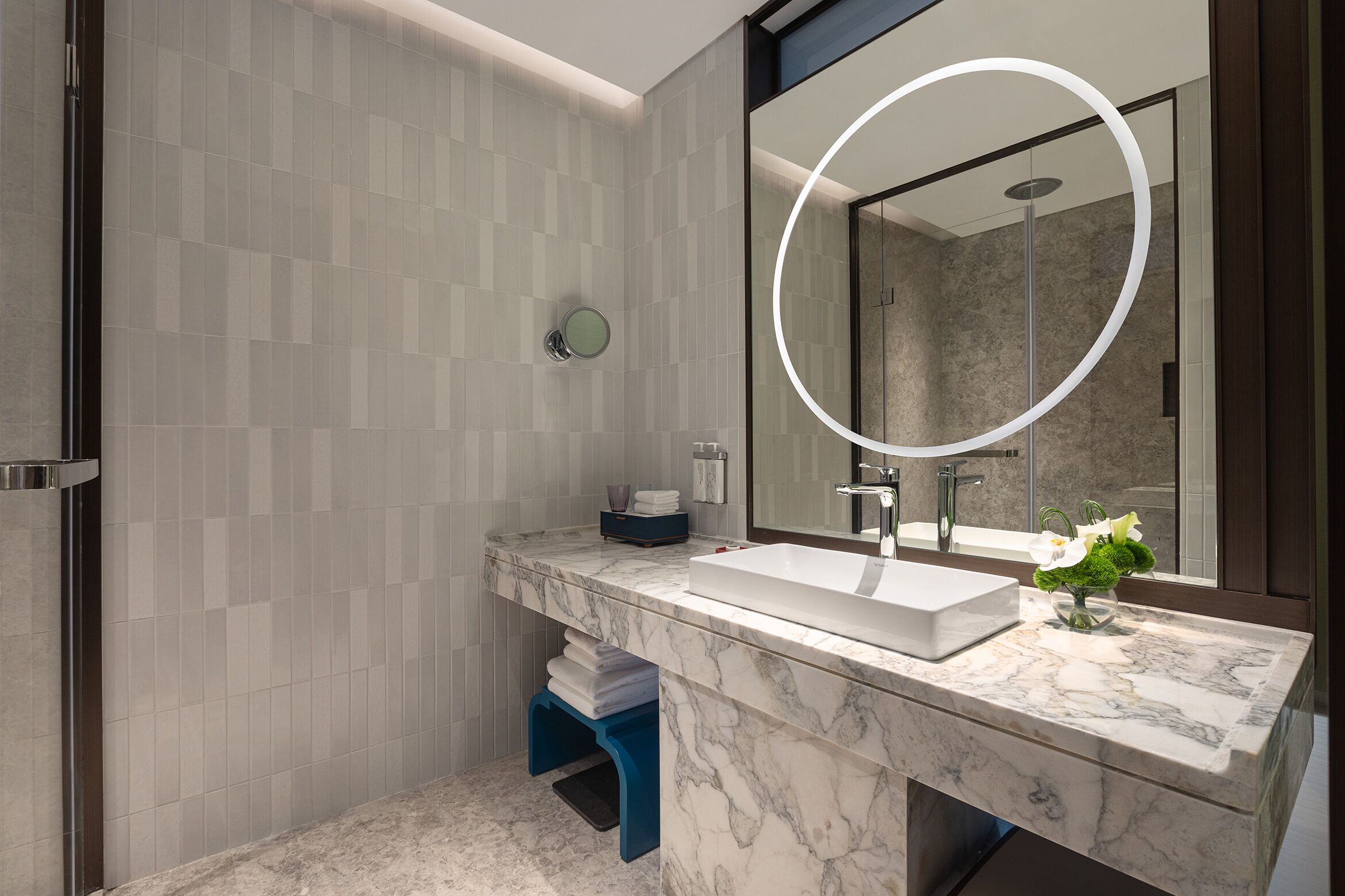A modern bathroom with a large mirror, marble countertop, and a shower visible in the reflection.