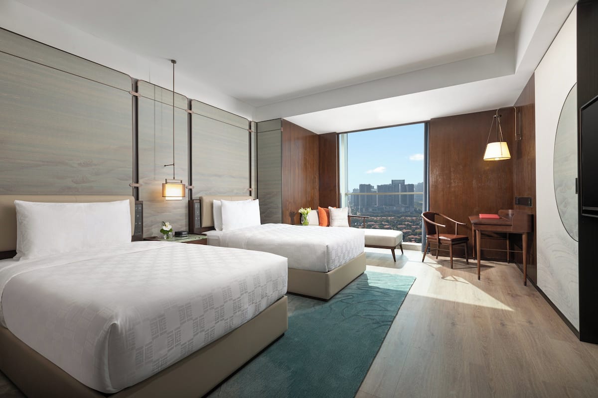 A modern hotel room with two beds, a desk, and a view of the city.