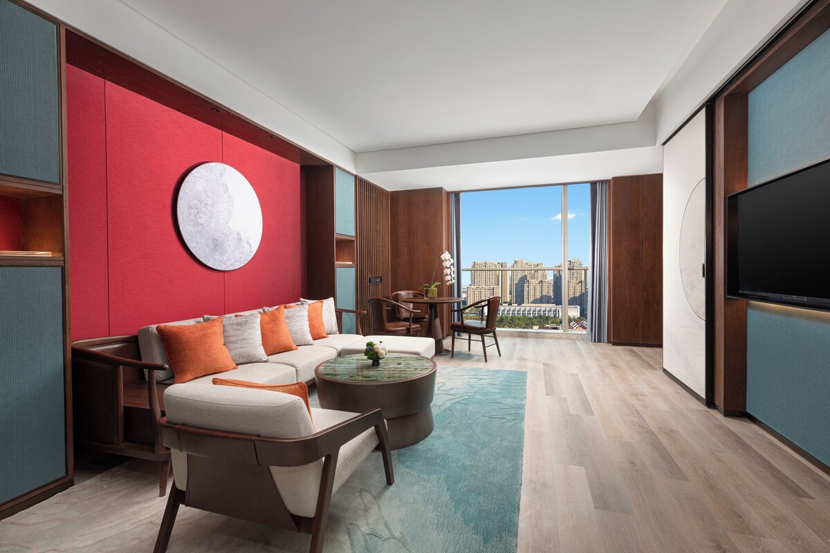A modern living room with a red accent wall, a round white artwork, a sofa with orange and gray cushions, a coffee table, and a TV on the right.