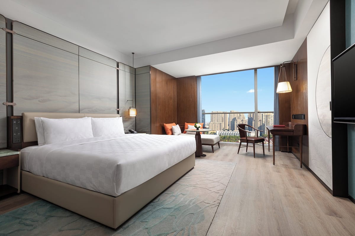 A modern hotel room with a large bed, a desk, a chair, and a view of the city.