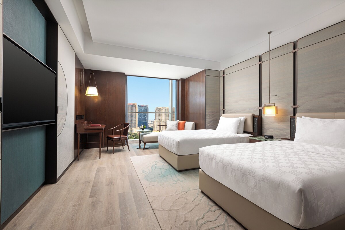 A hotel room with two beds, a desk, and a chair. There is a large window with a city view.