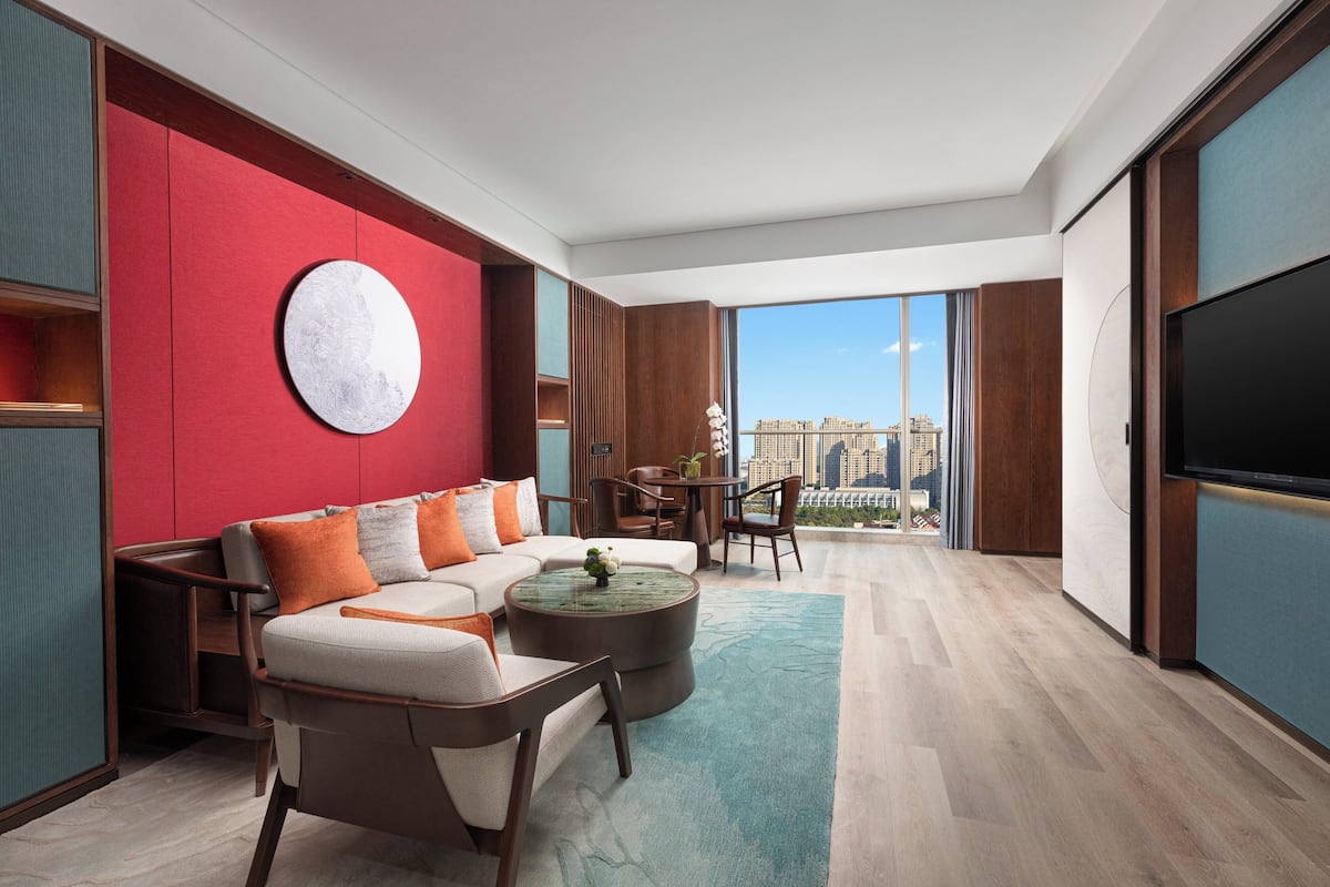 A modern living room with a red accent wall, a round white artwork, a sofa with orange and gray cushions, a coffee table, and a TV on the right.