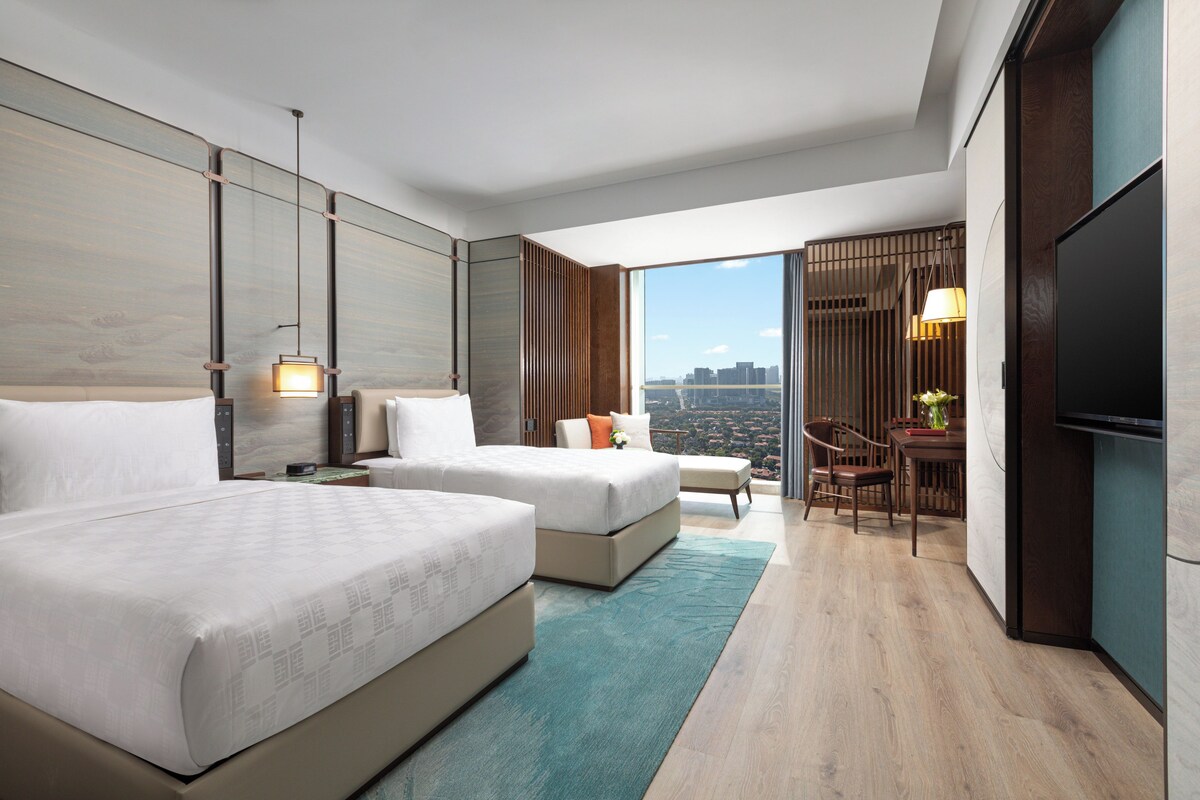A modern hotel room with two beds, a desk, a chair, and a large TV.