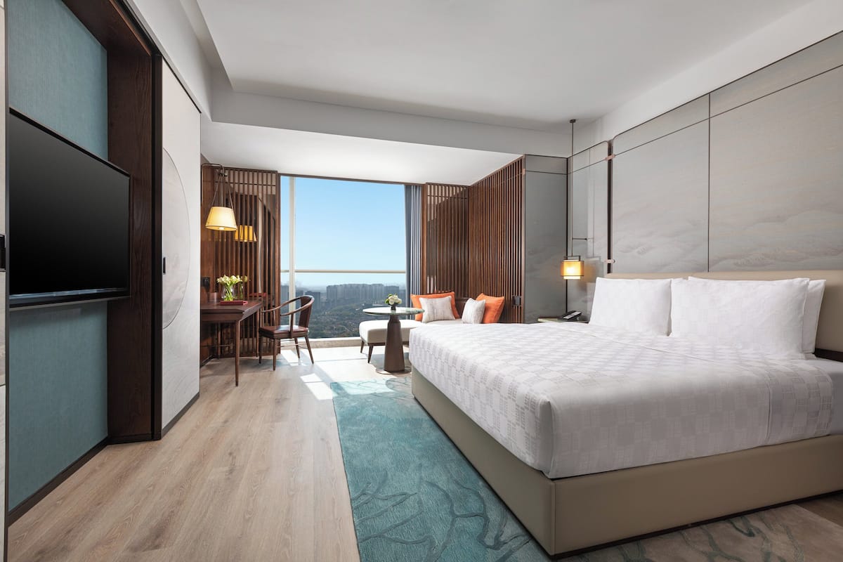 A modern hotel room with a large bed, a flat-screen TV, a dining area with a table and chairs, and a view of the cityscape.