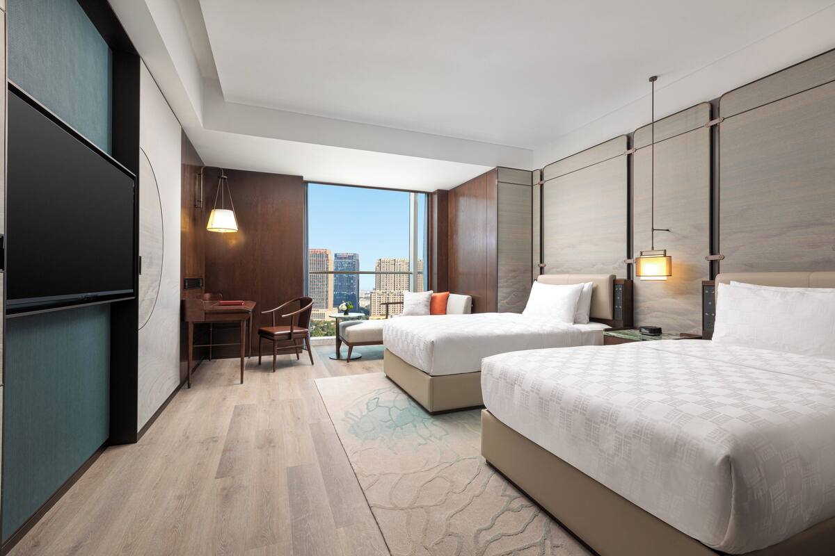A hotel room with two beds, a desk, and a chair. There is a large window with a city view.