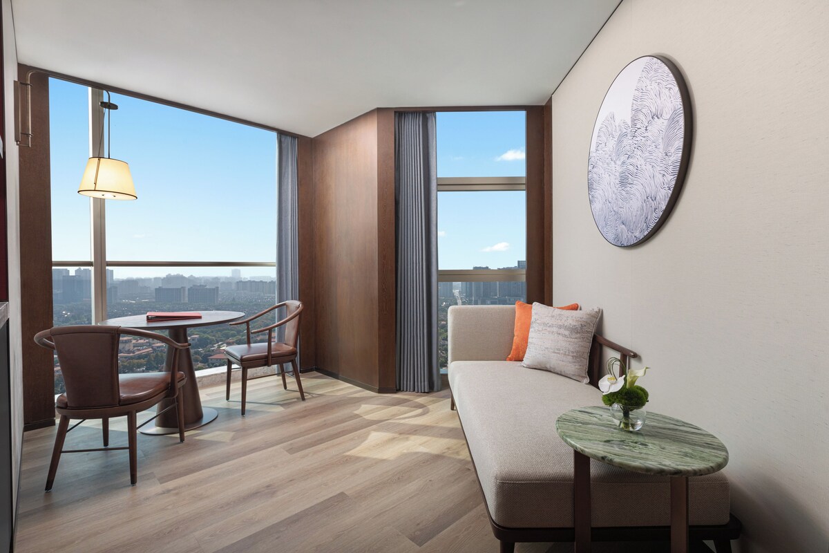 A modern hotel room with a sofa, a small table with a vase, a dining area with chairs, and large windows offering a city view.