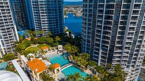 A high-rise hotel with a central swimming pool surrounded by palm trees and a view of the sea. - Stunning Chevron Apartment: Beach View| Pools| 2BR (Surfers Paradise)
