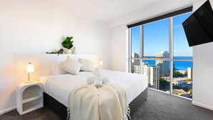 A hotel room with a large bed, a nightstand, a lamp, a plant, and a view of the city and ocean. - Stunning Chevron Apartment: Beach View| Pools| 2BR (Surfers Paradise)