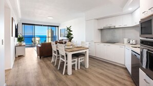 A modern kitchen with white cabinets, a dining area with a wooden table and chairs, and a view of the ocean through large windows. - Stunning Chevron Apartment: Beach View| Pools| 2BR (Surfers Paradise)