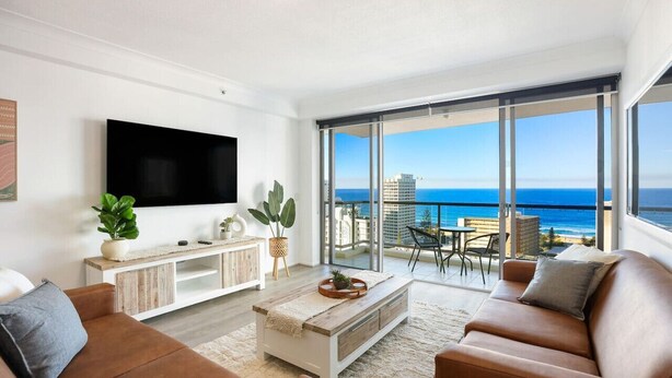 A modern living room with a large TV, a wooden coffee table, and a sofa. There is a balcony with a view of the ocean and buildings. - Stunning Chevron Apartment: Beach View| Pools| 2BR (Surfers Paradise)