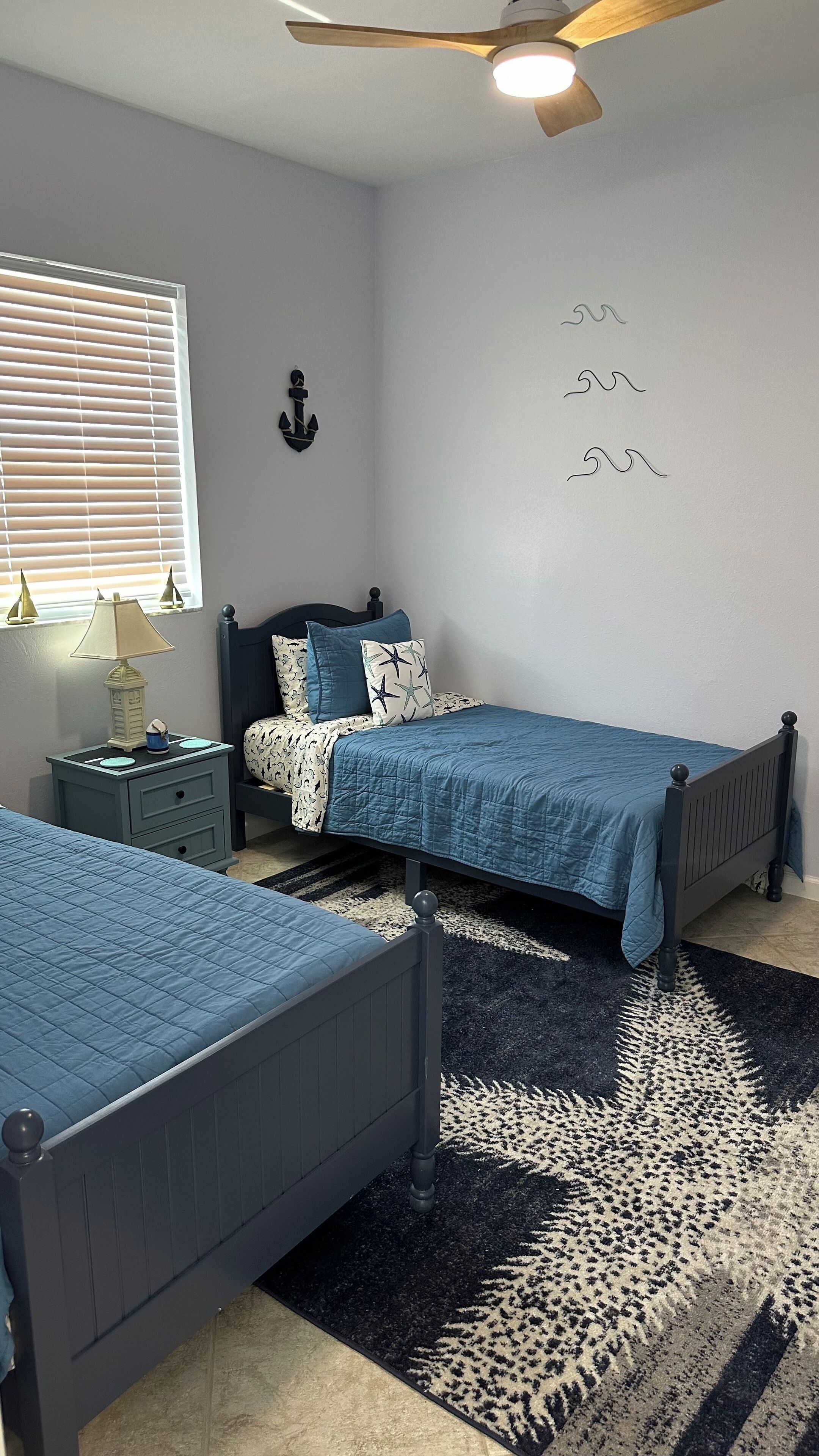 Une chambre avec deux lits simples, une table de chevet avec une lampe, un ventilateur de plafond et un mur orné d’un motif décoratif représentant une ancre et des étoiles.