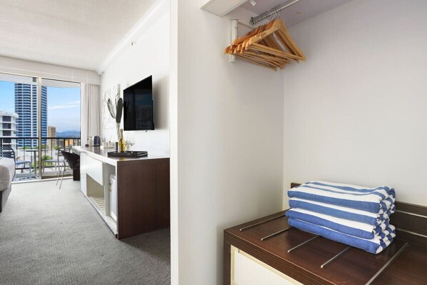 A hotel room with a balcony, a desk with a flat-screen TV, and a stack of towels on a wooden dresser.
