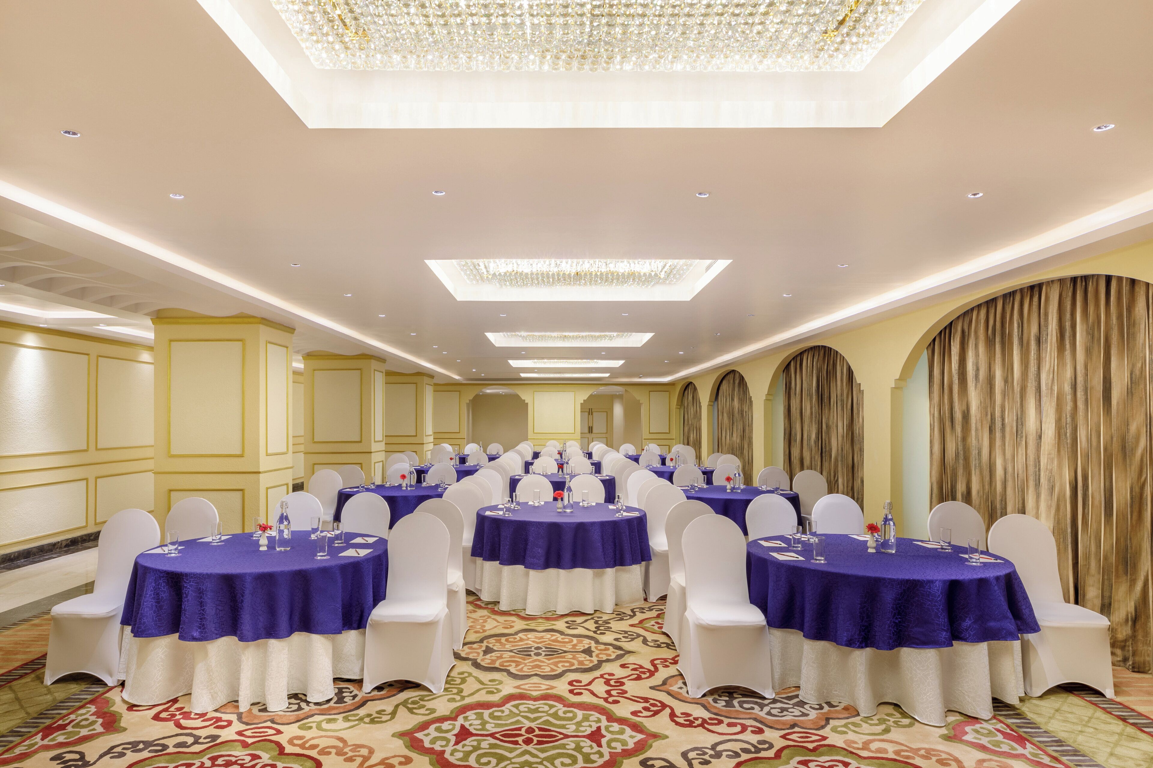 A banquet hall with round tables covered in blue tablecloths, white chairs, and a patterned carpet.