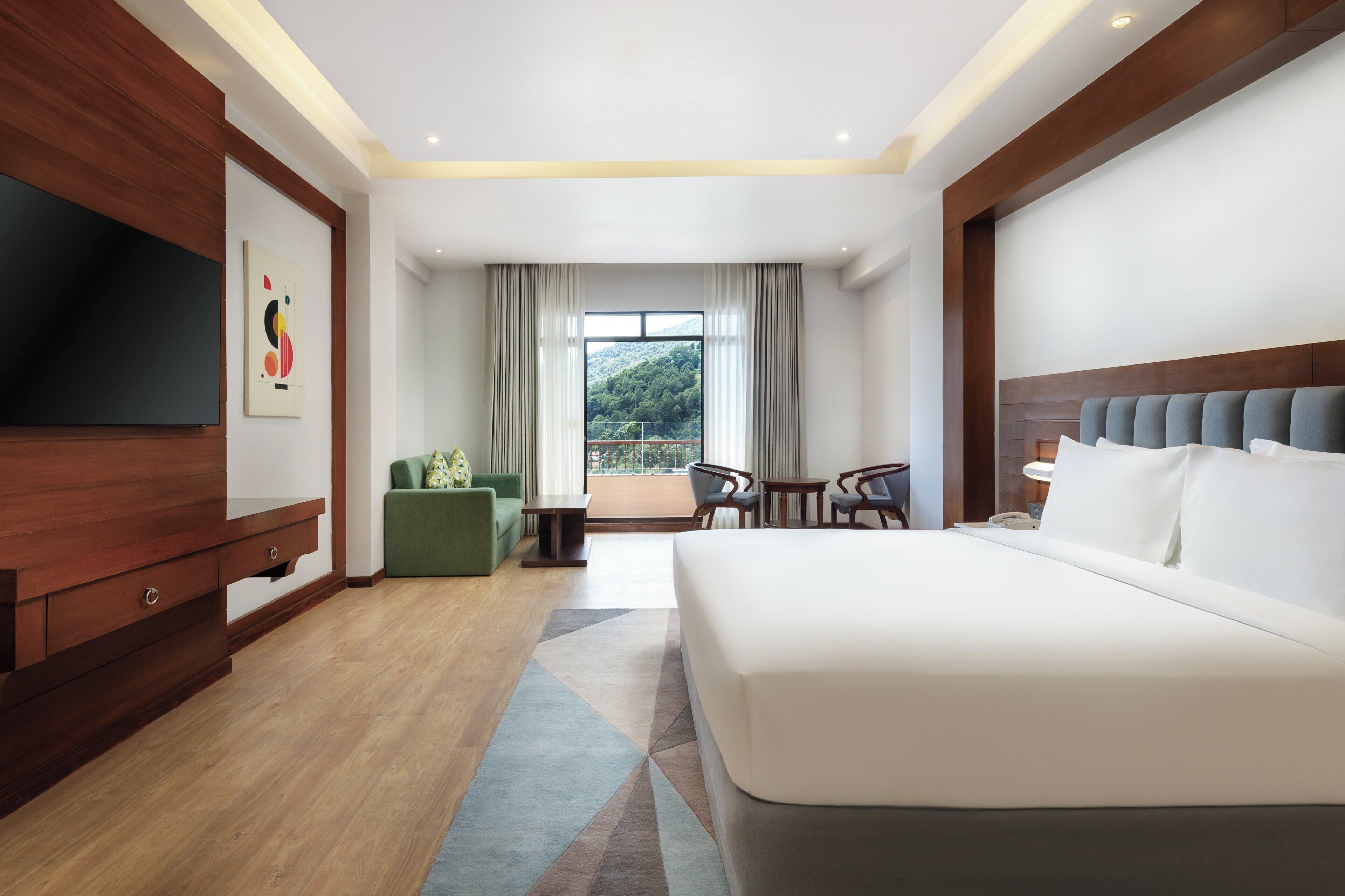 A modern hotel room with a large bed, a TV, a green sofa, a small table, and a balcony with a view of greenery.