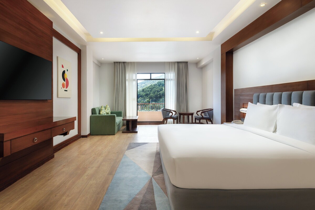 Une chambre d’hôtel moderne avec un grand lit, une télévision, un canapé vert, une petite table et un balcon donnant sur un paysage verdoyant.