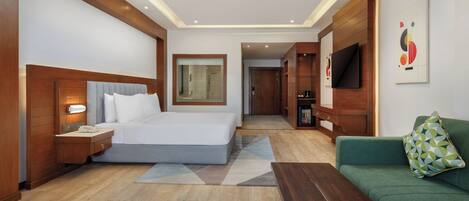 A modern hotel room with a large bed, a green sofa, a wooden coffee table, a TV, and a small kitchen area.