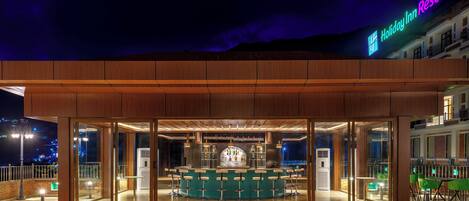 Una zona de bar con mostrador y taburetes, un ventanal grande y vistas nocturnas de la ciudad.
