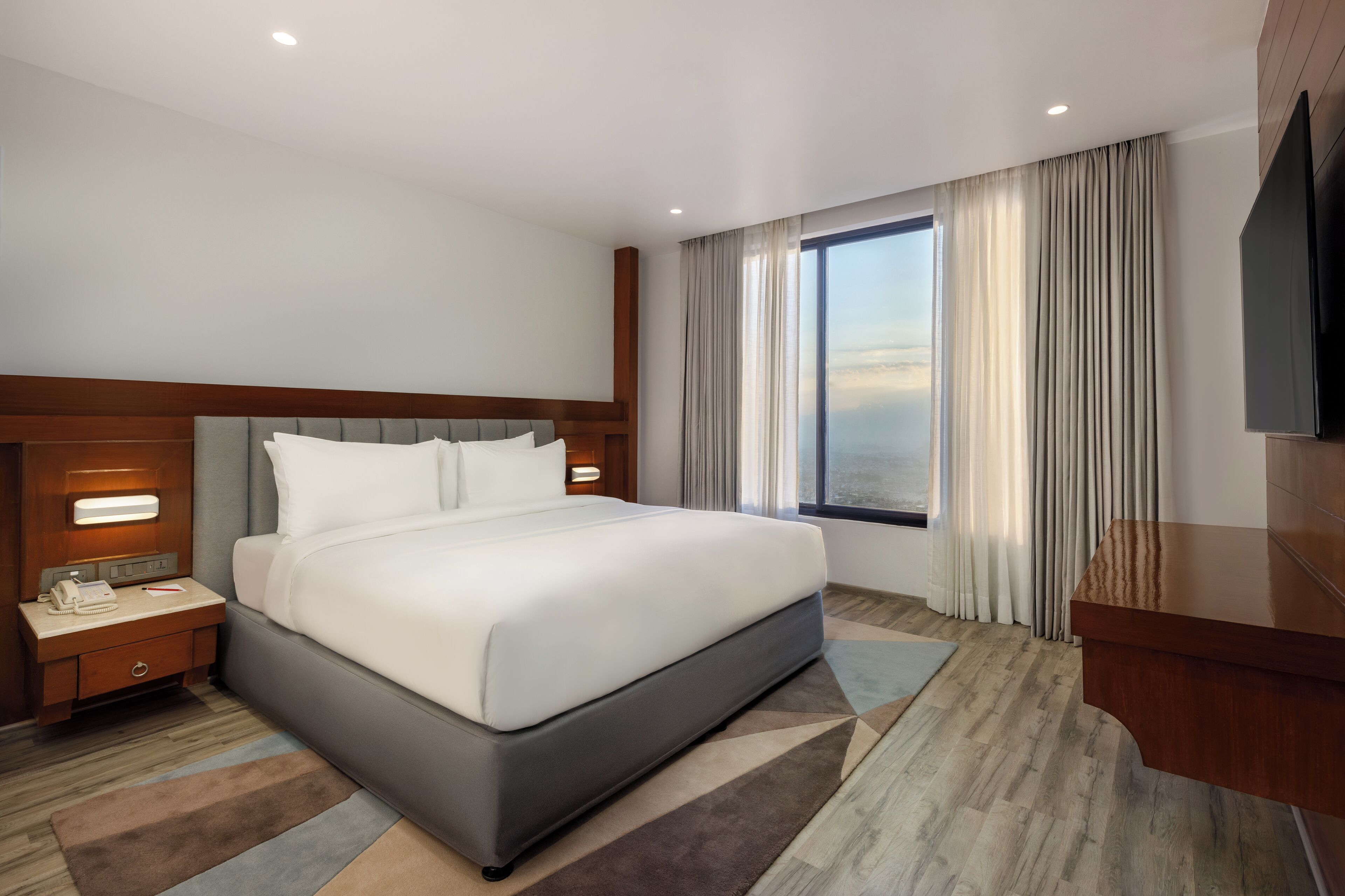 Un hotel moderno con una cama grande, una mesita de noche con teléfono y lámpara, un televisor y vistas a la ciudad a través de amplios ventanales.