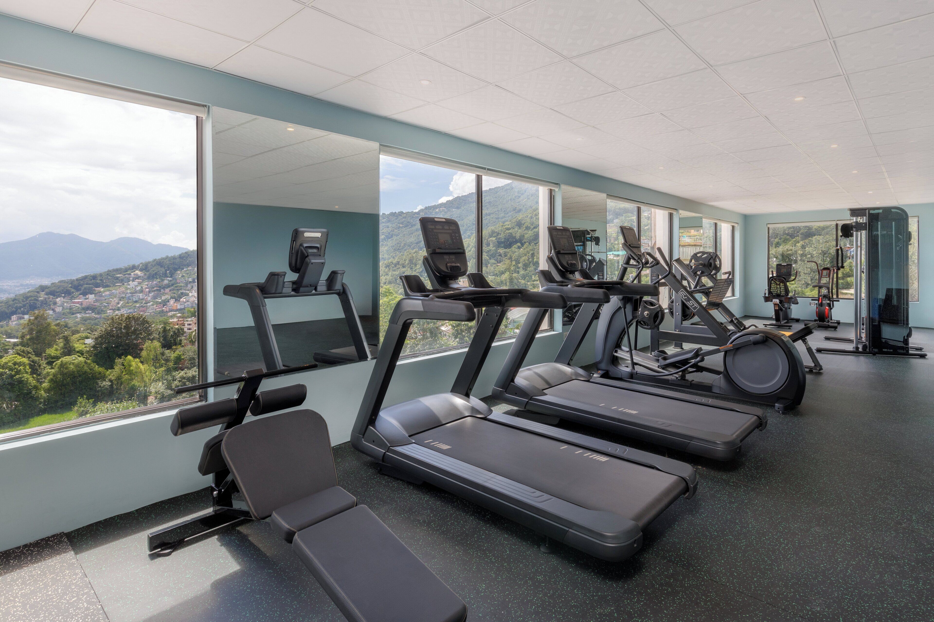 Un gimnasio moderno con cintas de correr, máquinas de pesas y una zona de bancos, con amplias ventanas y vistas a la montaña.