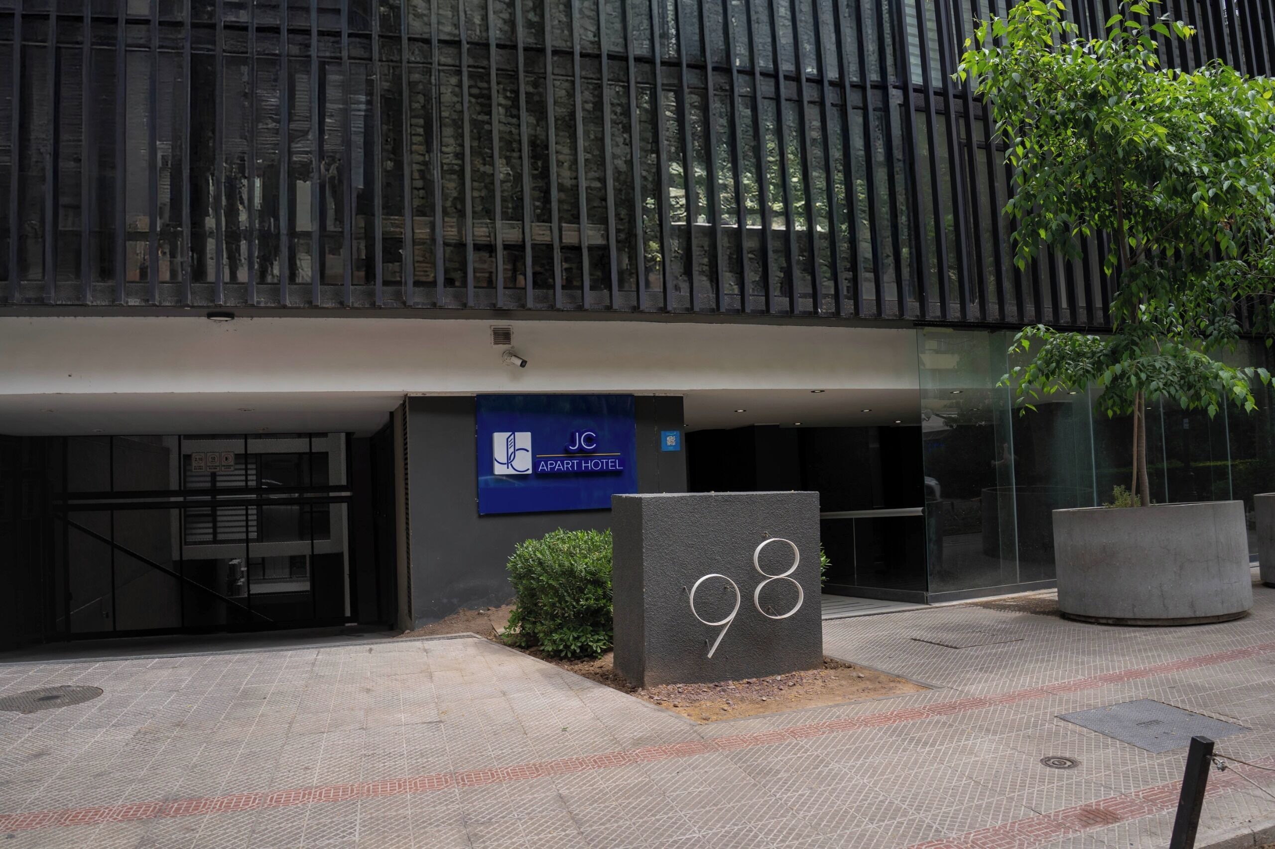 Um edifício moderno com uma placa indicando JC Apartment Hotel, um vaso grande com uma árvore e uma placa com o número 98 no chão.
