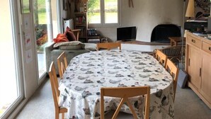 Un comedor con una mesa redonda cubierta por un mantel estampado con pájaros, rodeada de sillas de madera. Se ve una ventana con vistas a la vegetación, y un ventilador de techo en lo alto.
