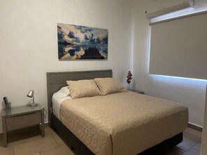 A bedroom with a bed, a nightstand, a lamp, a painting, and a window with blinds.