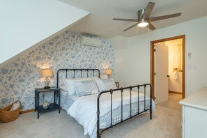 A bedroom with a sloped ceiling, a bed with a metal frame, two bedside tables with lamps, a wall-mounted air conditioner, and a door leading to another room. - Cozy Cabin in town with a View! Hot Tub+Grill+WiFi (Burnsville)